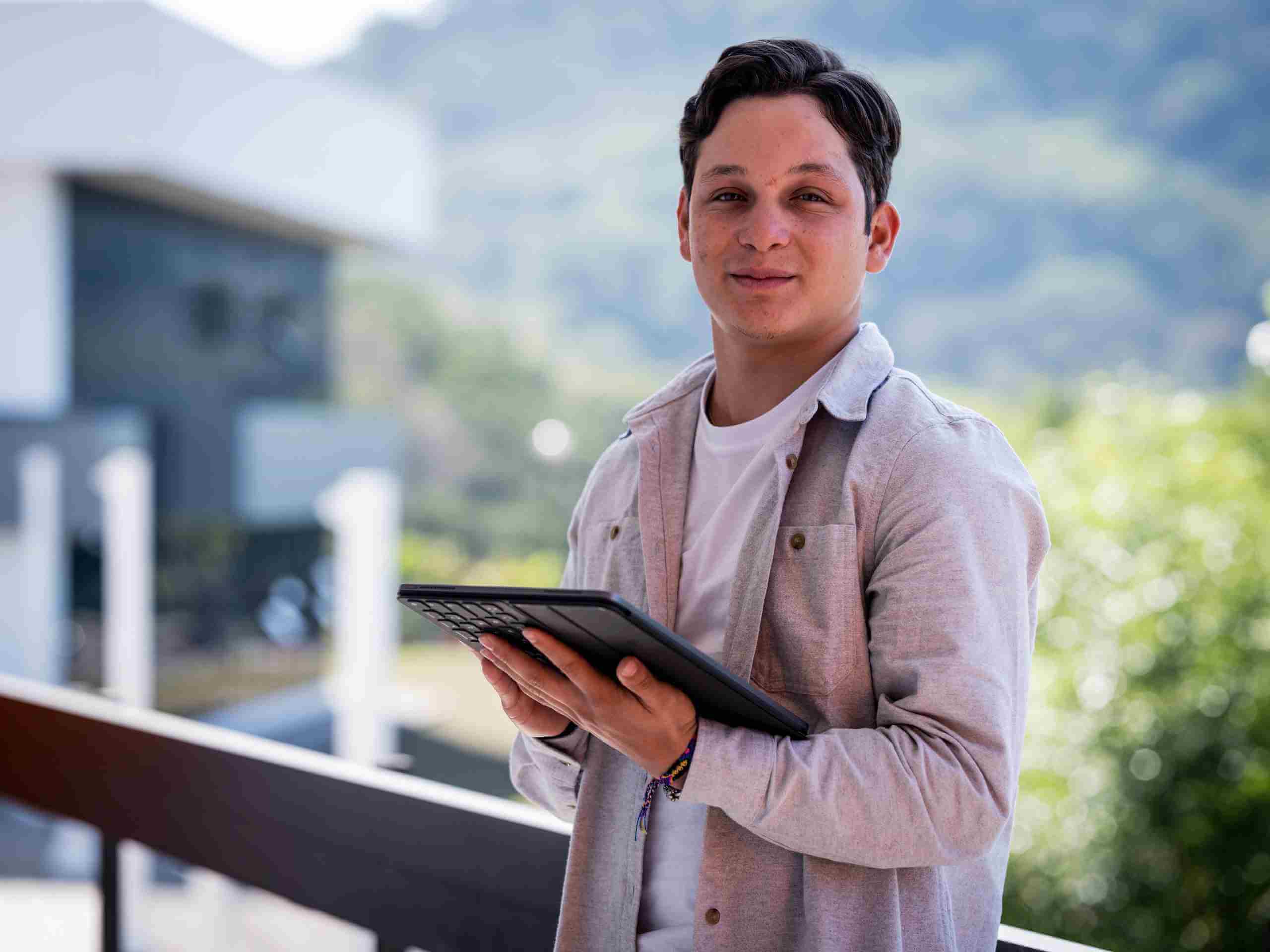 Joven con camisa gris claro sostiene una tablet mientras posa sonriente en una terraza con fondo de vegetación y edificios modernos.